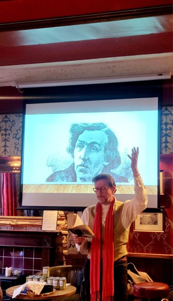 A speaker reading poetry on stage, with a large illustration of William McGonagall projected behind him, in a cozy pub setting.
