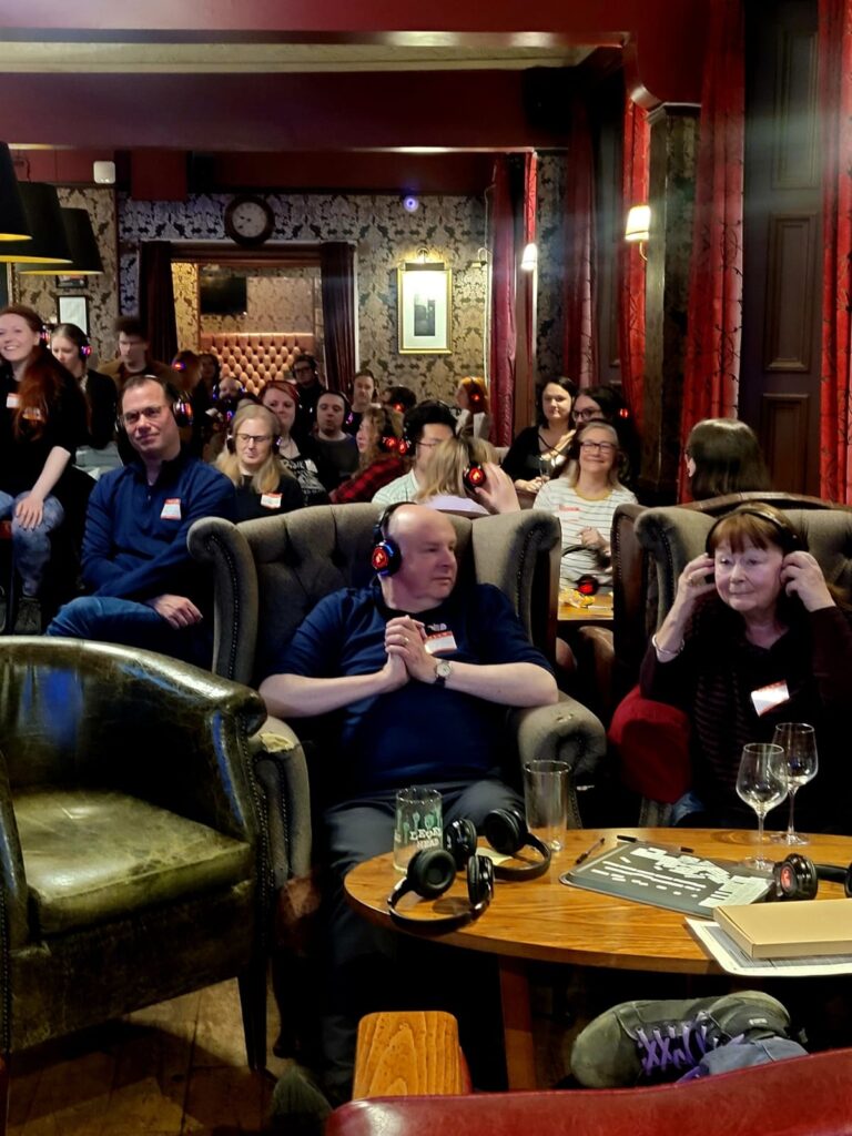 A group of people seated in a cozy pub, with some wearing headphones and engaging in an event. The background features decorative wallpaper and dim lighting, creating a warm atmosphere.