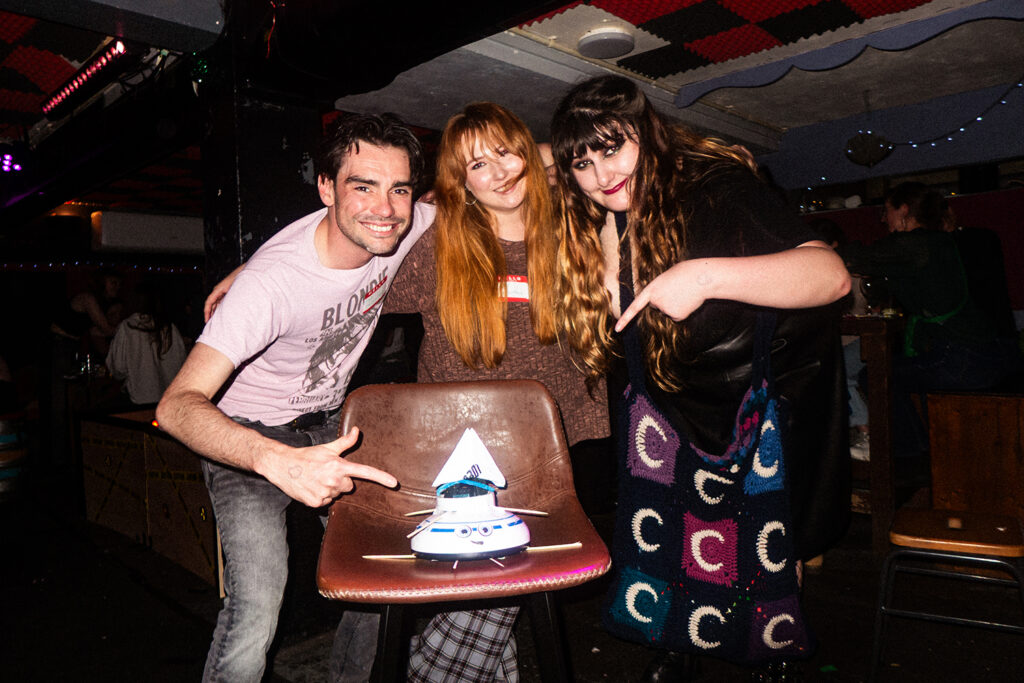 Three participants posing with a customized robot vacuum on a chair at a lively event, showcasing excitement and creativity.