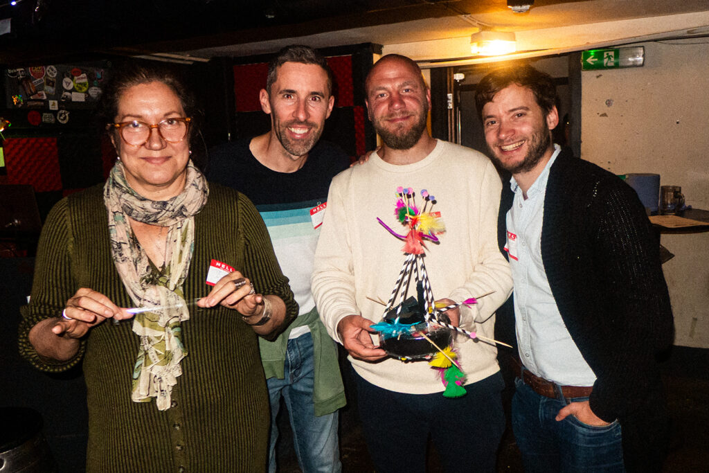 A group of four people stands together in a lively venue, each holding crafting materials. The woman on the left has glasses and wears a green sweater, while the others, including two men and another woman, smile and pose with a colorful, creatively decorated robot vacuum. The background features walls adorned with stickers and soft lighting, capturing the fun atmosphere of the event.