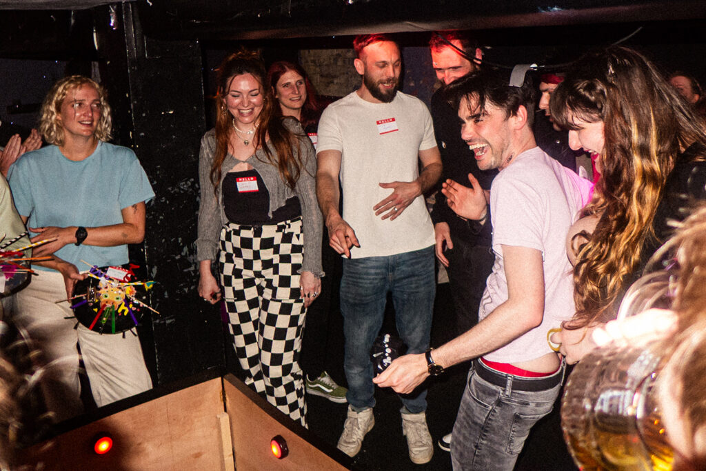 A lively scene from the Robot Vacuum Fight Club, showcasing enthusiastic participants cheering and interacting in a playful atmosphere within a dimly lit venue.