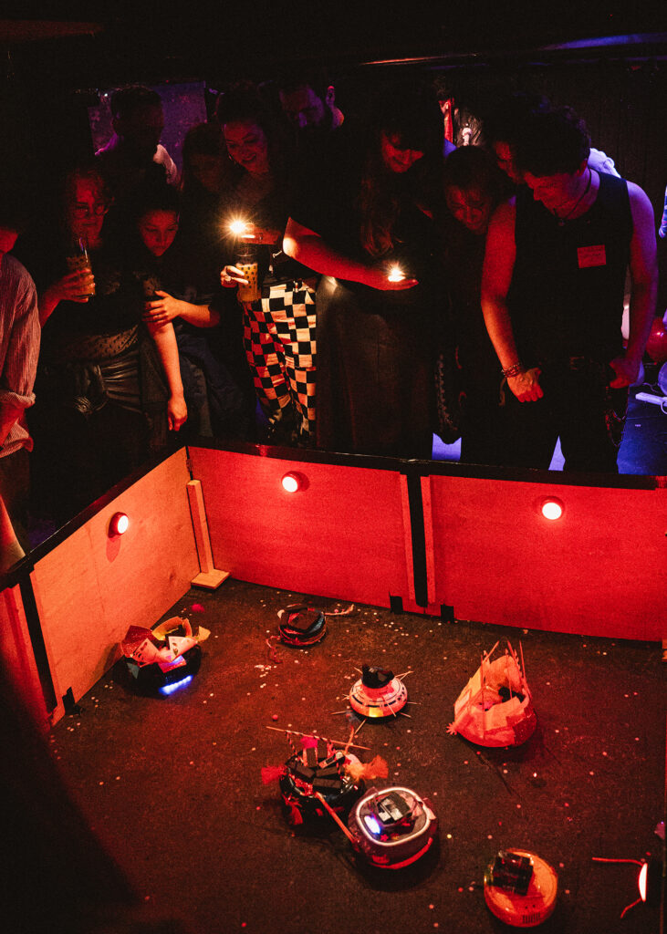 A lively scene at a robot vacuum competition, showcasing several modified robot vacuums in an arena surrounded by spectators holding lights, creating a vibrant atmosphere.