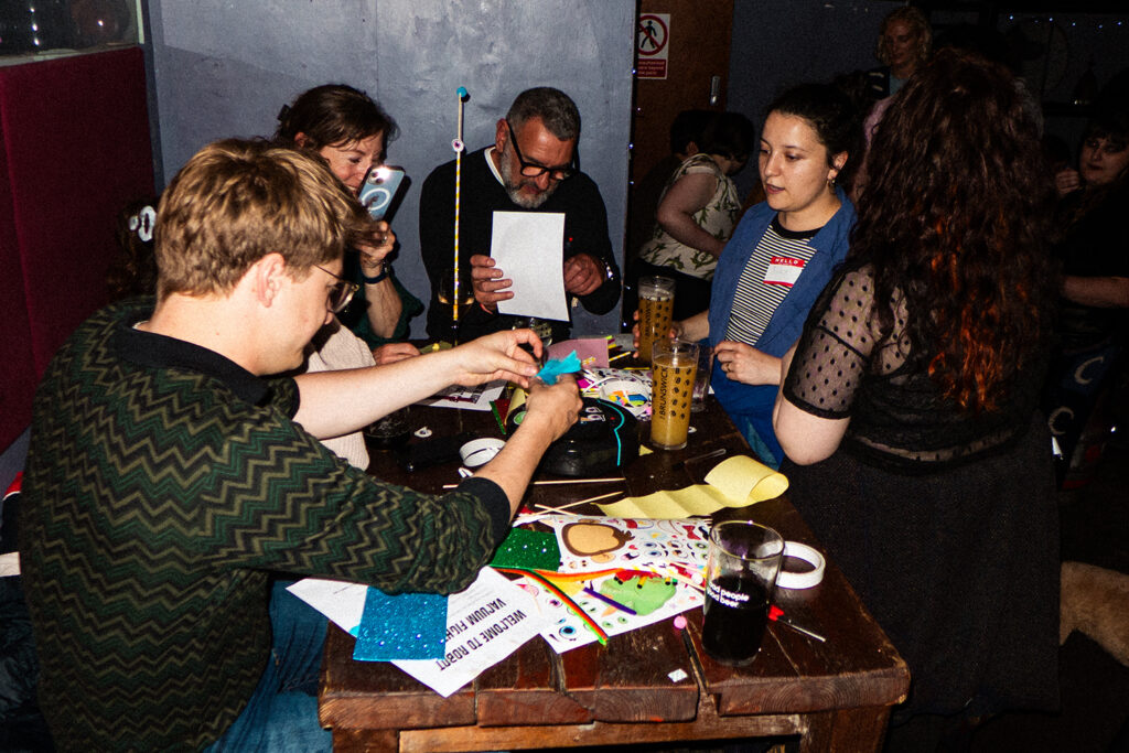 A group of people gathered around a table engaged in crafting activities with various materials, including colorful paper and drinks, in a lively setting.