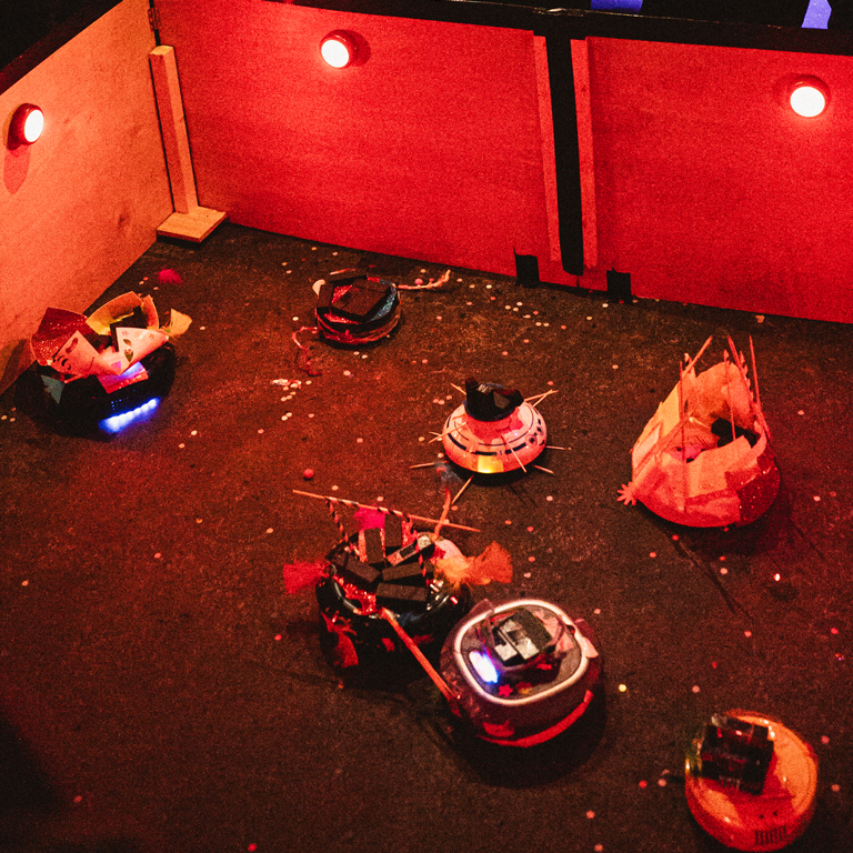 A colorful arena with several customized robot vacuum cleaners, each adorned with various decorations, competing in a lively and chaotic setting.